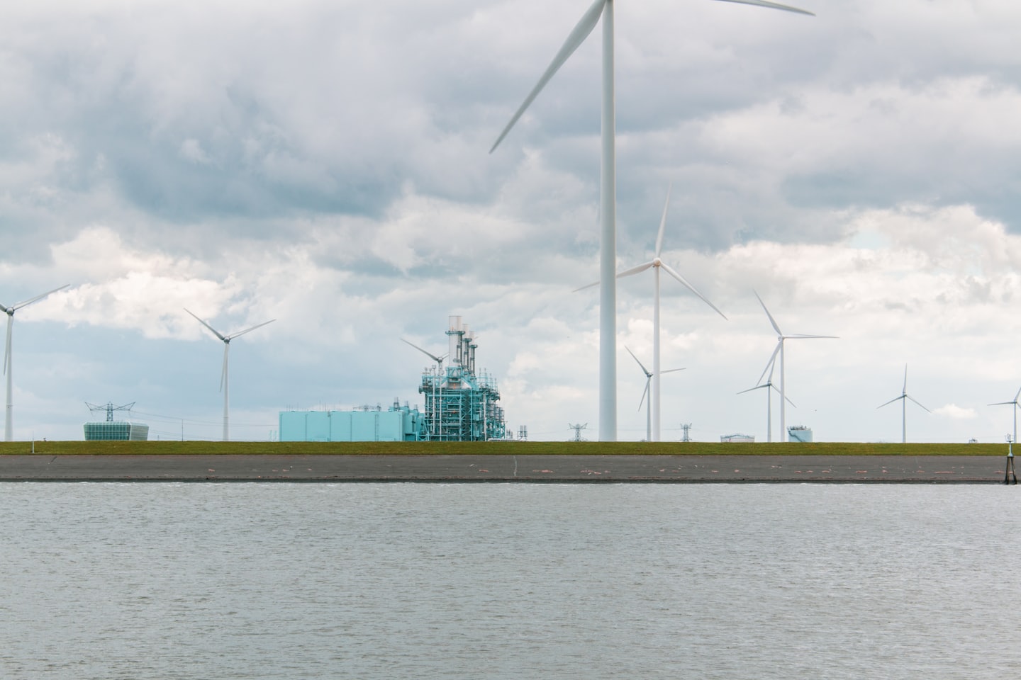 Turbine eoliche e impianto industriale su un argine erboso vicino all'acqua.